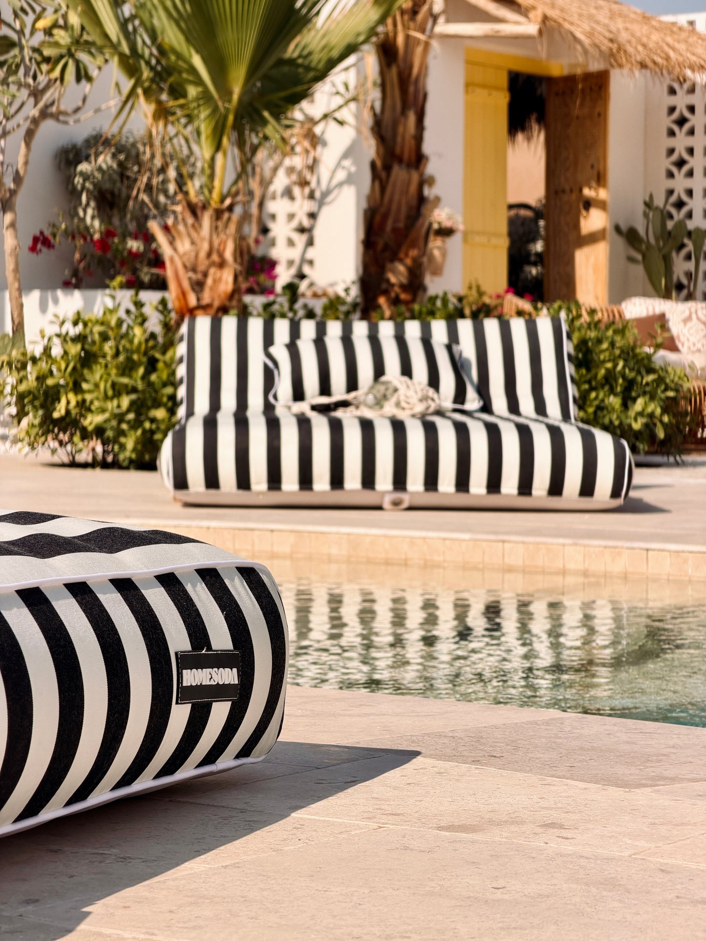 Black and white striped outdoor furniture by a poolside with palm trees and a building in the background.