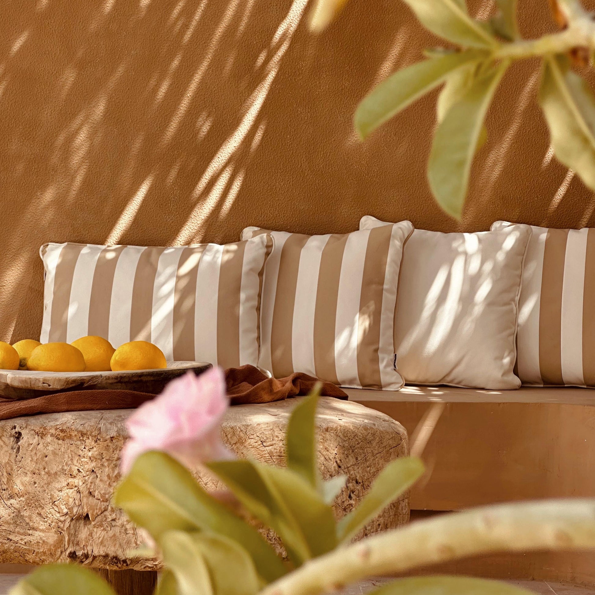 Beige outdoor cushion with striped cushions against a warm-toned wall, surrounded by plants.
